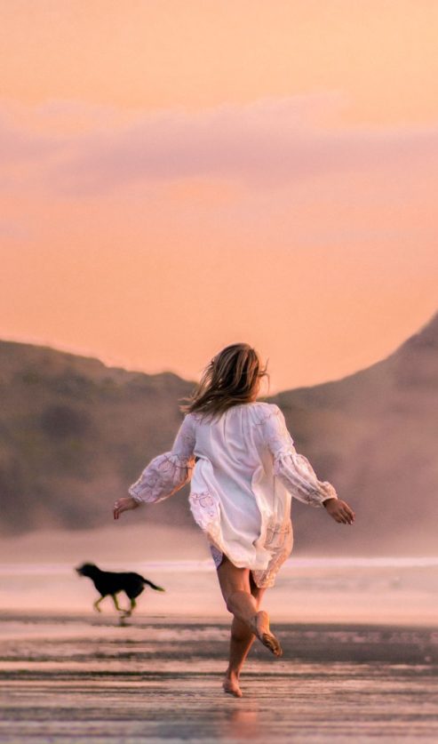 Žena bosonoga trči plažom u suton dok se u pozadini uzdižu litice, simbolizirajući nove početke i promjene koje donosi mjesečni horoskop za kolovoz 2026