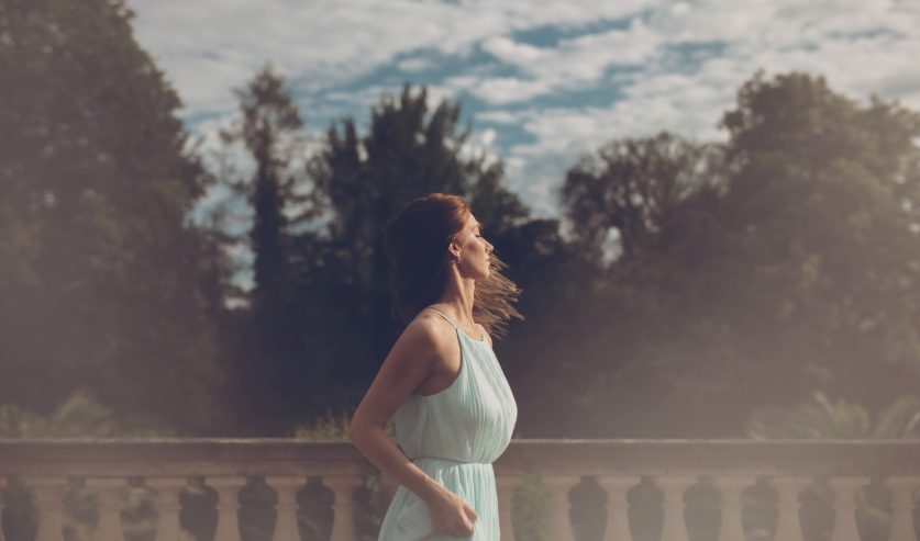 Žena u haljini stoji na elegantnoj terasi okrenuta prema svjetlu, simbolizirajući skl